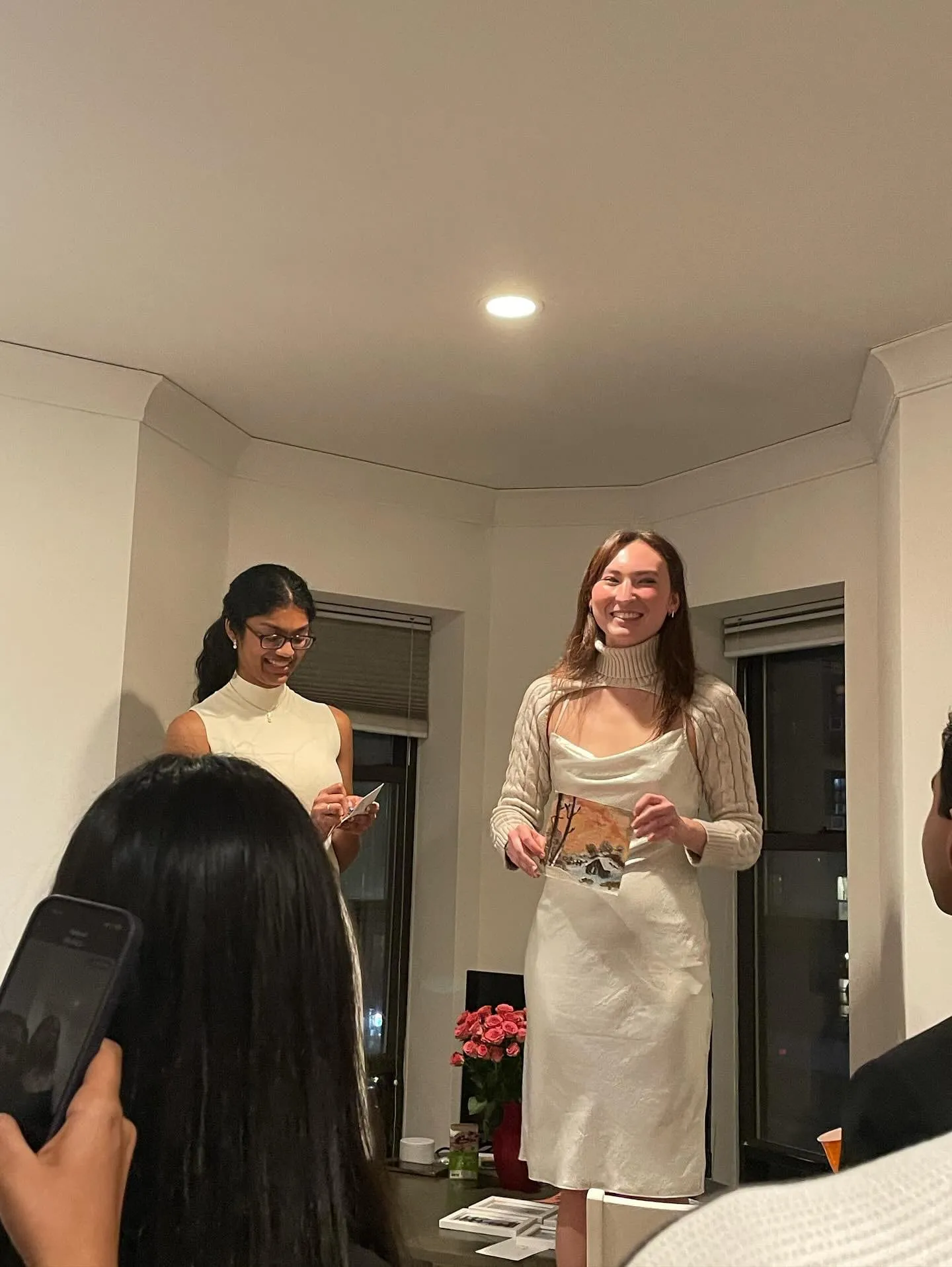 sahi and a co-host in white, smiling as a small painting is auctioned off in a living room